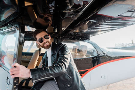 cheerful pilot in stylish sunglasses and leather jacket opening door in helicopterの写真素材