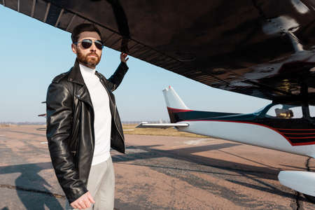 bearded pilot in sunglasses and leather jacket standing under aircraft wingの写真素材