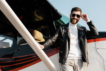 cheerful bearded man adjusting sunglasses near helicopter