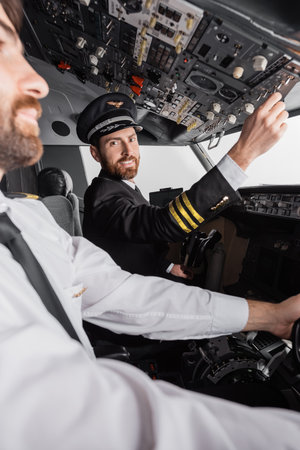 cheerful captain in cap and uniform reaching overhead panel near co-pilot in airplane simulatorの写真素材
