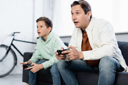 KYIV, UKRAINE - OCTOBER 11, 2021: Man playing video game with preteen son on couch at home