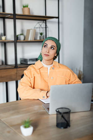 dreamy muslim woman looking away while sitting near laptop at homeの写真素材