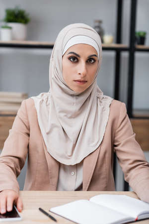 muslim woman in hijab sitting near blank notebook and mobile phone at homeの写真素材