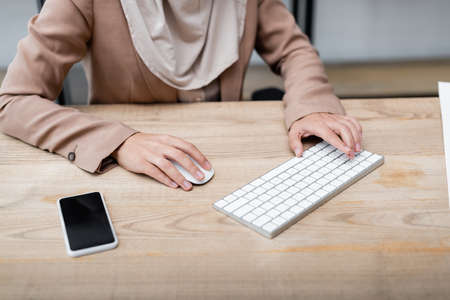 cropped view of woman using keyboard and computer mouse while working near smartphone with blank...