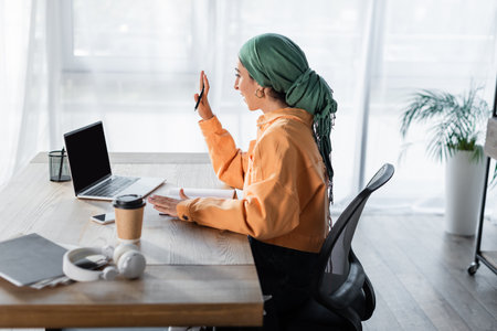 side view of muslim woman waving hand during video call on laptop