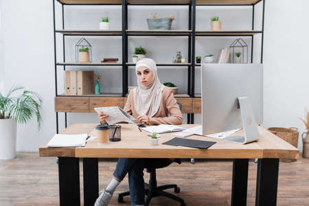muslim woman holding newspaper and looking at camera near computer on work desk at homeの写真素材
