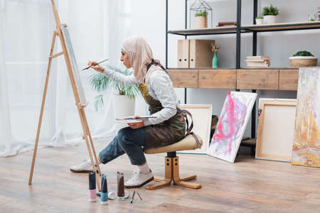 side view of muslim woman sitting on chair and drawing on easel at homeの写真素材