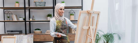 happy woman in hijab and apron drawing on easel at home, bannerの写真素材