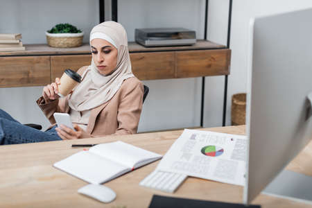 muslim woman with coffee to go and smartphone sitting near blank notebook and charts on deskの写真素材