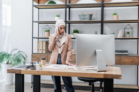 muslim woman standing near computer and documents on work desk while talking on mobile phoneの写真素材