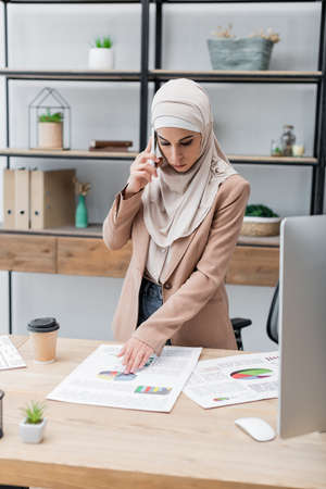 muslim woman talking on mobile phone near infographics and computer on deskの写真素材