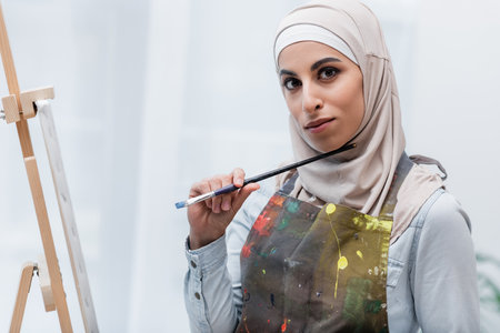 young muslim woman holding paintbrush while looking at camera near easelの写真素材