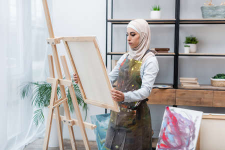 muslim woman in hijab and apron holding canvas near easel at homeの写真素材