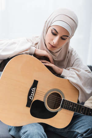 young arabian woman in hijab leaning on acoustic guitar while sitting at homeの写真素材