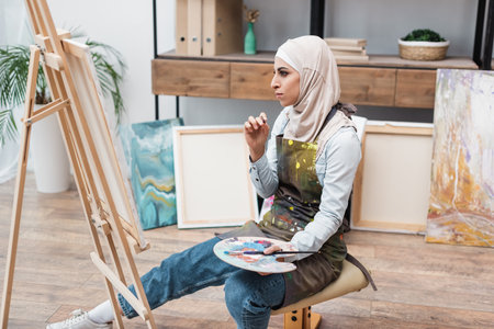 muslim woman with palette sitting near easel in home studioの写真素材