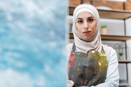 young arabian woman in hijab and apron looking at camera near blurred painting