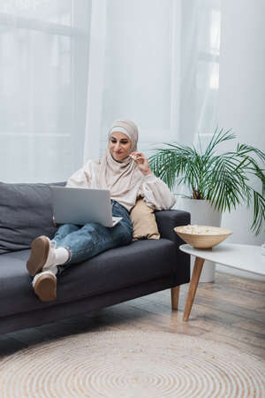 cheerful arabian woman in hijab eating popcorn and watching film on laptop on couchの写真素材