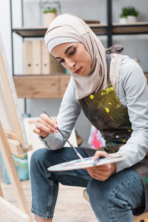young muslim woman with palette and paintbrush sitting near blurred easelの写真素材