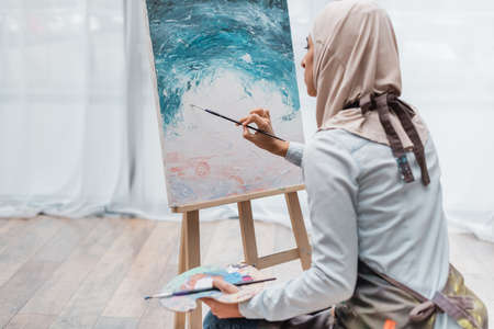 young muslim woman in hijab painting on easel at homeの写真素材