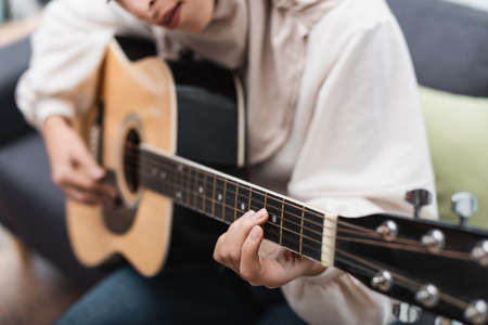 cropped view of blurred muslim woman playing acoustic guitar at home