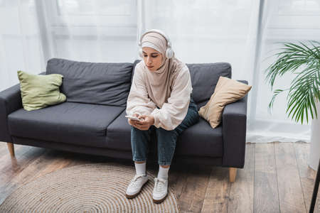 young muslim woman sitting on couch with smartphone and listening to music in headphonesの写真素材