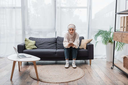 muslim woman in headphones using smartphone while listening to music in living roomの写真素材