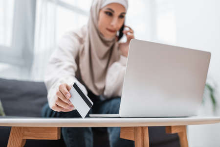 blurred arabian woman talking on mobile phone while holding credit card near laptopの写真素材