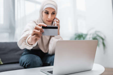 selective focus of credit card in hand of blurred arabian woman calling on smartphone near laptopの写真素材