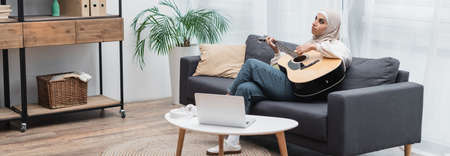 young muslim woman playing guitar while sitting on couch near laptop in living room, bannerの写真素材