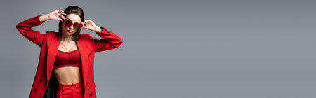 young woman in red suit with crop top and trendy sunglasses looking at camera isolated on dark grey, bannerの写真素材