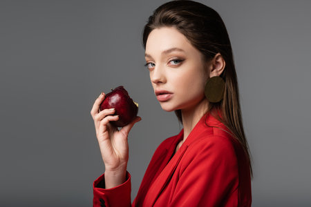 stylish young woman in red blazer and earring holding apple isolated on grayの写真素材