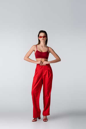 full length of young woman in sunglasses and red outfit posing on grayの写真素材
