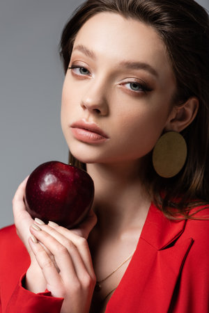 pretty woman in red blazer and earring holding ripe apple isolated on grayの写真素材