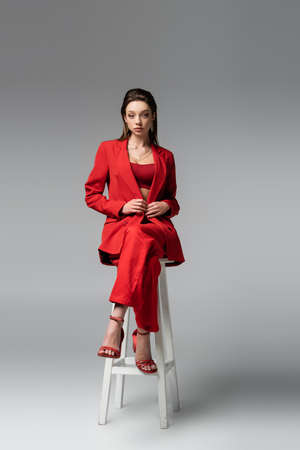 full length of pretty woman in red suit and shoes sitting on white chair and looking at camera on dark grayの写真素材