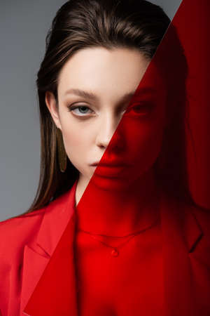 trendy young woman looking at camera through red glass isolated on grayの写真素材