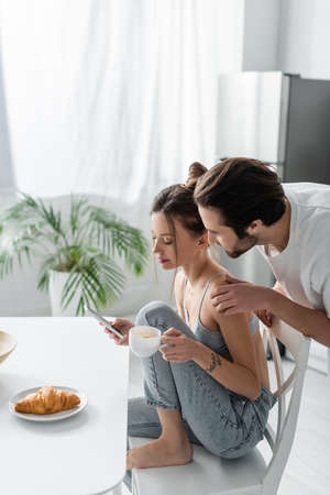 young woman in bra and jeans using smartphone and holding cup near boyfriend in kitchenの写真素材
