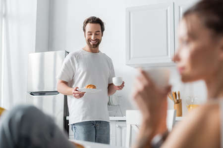 happy bearded man holding plate with croissant and cup near blurred woman on foregroundの写真素材