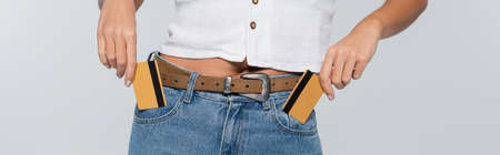 cropped view of young woman putting credit cards with cashback in pockets isolated on grey, banner