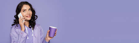 happy young woman in raincoat holding paper cup and talking on smartphone isolated on purple, bannerの写真素材