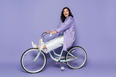 full length of amazed young woman in violet raincoat riding bicycle on purpleの写真素材