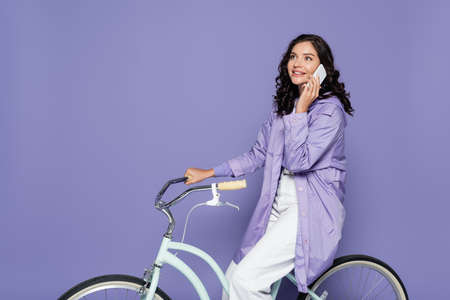 happy young woman in violet raincoat riding bicycle and talking on smartphone isolated on purple