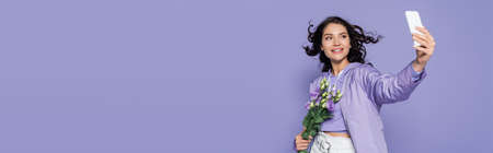 happy young woman holding flowers and taking selfie on smartphone isolated on purple, banner