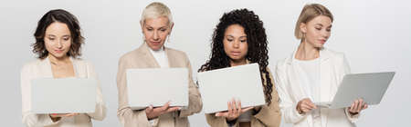 Interracial businesswomen in formal wear using laptops isolated on grey, banner