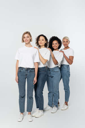 Full length of multiethnic women in jeans and white t-shirts looking at camera on grey background