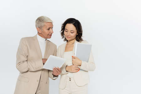 Senior businesswoman holding digital tablet near colleague with paper folder isolated on greyの写真素材