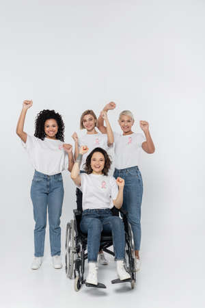 Smiling interracial women with pink ribbons showing yes gesture near friend in wheelchair on grey backgroundの写真素材