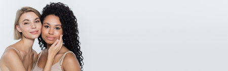 Blonde woman touching face of african american friend isolated on grey, bannerの写真素材