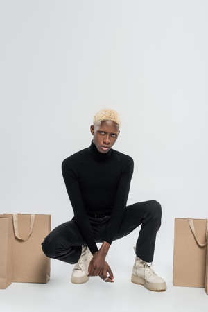 full length of young african american man sitting with paper bags on grey