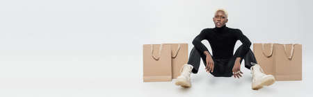 full length of blonde african american man sitting near paper bags on grey, bannerの写真素材