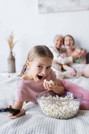 Excited girl holding popcorn and remote controller near blurred mothers on bedの写真素材